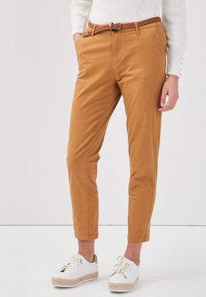 Femme portant un pantalon court de couleur camel avec une ceinture tressée, un haut blanc à manches longues et des baskets blanches à lacets, sur un fond uni.