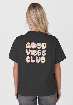 Osoba blond o odwróconych plecach, ubrana w czarną koszulkę z tekstem „GOOD VIBES CLUB” oraz trzema uśmiechniętymi buźkami w pastelowych kolorach na plecach.