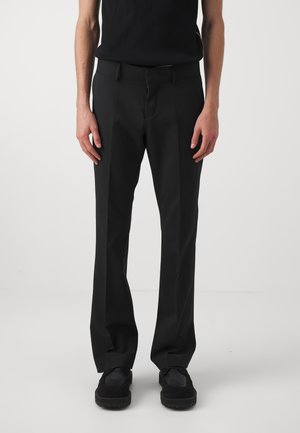 Mężczyzna w dopasowanych czarnych spodniach, czarnej koszulce bez rękawów i czarnych butach, stojący na jasnym, jednolitym tle.