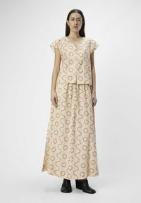 Conjunto de dos piezas que incluye una blusa de manga corta y escote en V, y una falda maxi. Tejido beige claro con un patrón de líneas onduladas rojas y puntos. Botas negras hasta el tobillo.