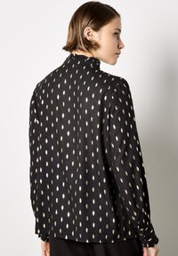 Black blouse featuring gold leaf patterns, long sleeves with gathered cuffs, and a ruffled collar. Smooth, lightweight fabric.