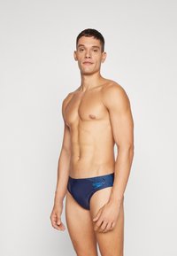 Homem sem camisa a usar um fato de banho azul marinho com logótipo azul claro, de pé contra um fundo cinza claro liso.