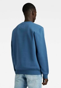 Hombre vistiendo un sudadera azul lisa y unos jeans azul claro, visto desde atrás sobre un fondo blanco.