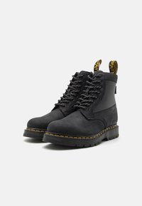 Botine negre cu șireturi, fabricate din suede, cu o textură netedă, având o talpă din cauciuc, cusături galbene și tab-uri întărite la călcâi.
