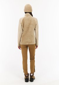Beige fleece jasje met een gestructureerde achterkant, gecombineerd met een paar bruine broeken en stevige laarzen. Een gebreide beige beanie maakt de outdoor outfit compleet.