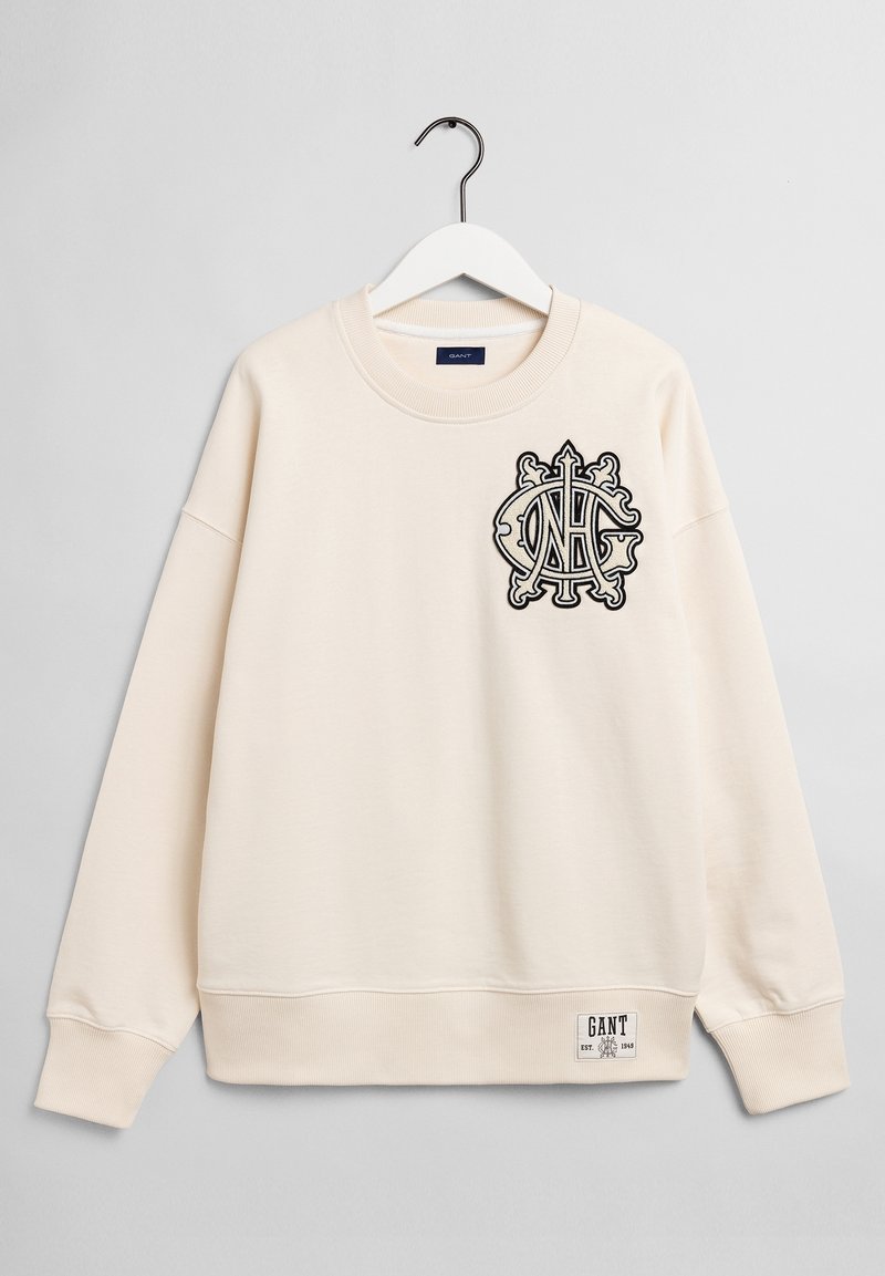 Sweatshirt crème à col rond, avec un logo noir brodé sur la poitrine et une étiquette tissée sur l'ourlet inférieur gauche.