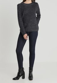 Dunkelgrauer, strukturierter Pullover mit rundem Ausschnitt, kombiniert mit dunklen Skinny-Jeans und glänzenden schwarzen Ankle-Boots mit Blockabsätzen.