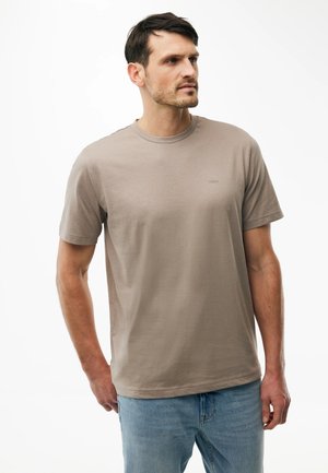 Hombre con cabello corto y oscuro, vestido con una camiseta beige lisa de manga corta y vaqueros azul claro, mirando hacia su izquierda.