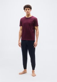 Νεαρός άνδρας στέκεται ξυπόλυτος, φορώντας μπορντό κοντομάνικο πουκάμισο και σκούρο μπλε παντελόνι φόρμας, σε απλό λευκό φόντο.