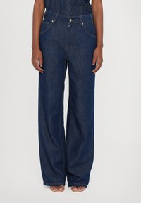 Calças de ganga largas de cor azul escuro, com bolsos frontais, fecho de botão e costuras visíveis.