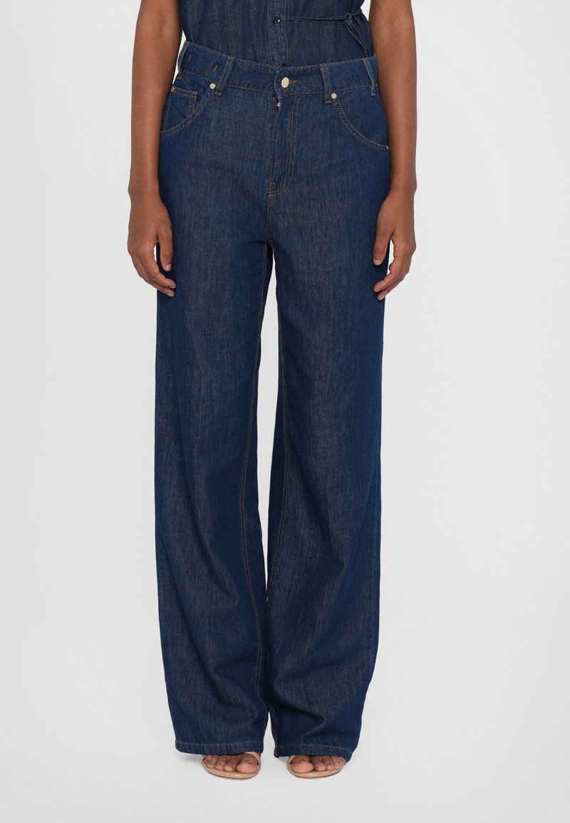 Calças de ganga largas de cor azul escuro, com bolsos frontais, fecho de botão e costuras visíveis.