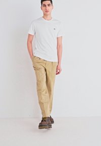 White crew-neck t-shirt paired with tan chinos and brown lace-up boots. Linen-like texture with minimal branding on the shirt.