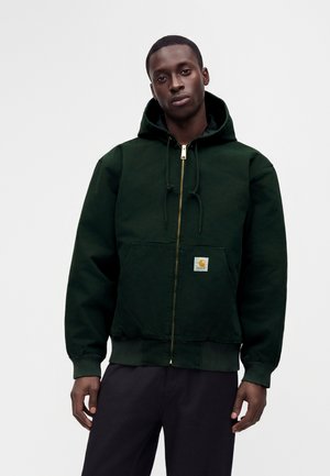 Jeune homme portant un sweat à capuche zippé vert foncé avec des poches avant et un petit patch avec le logo Carhartt, debout devant un fond uni.
