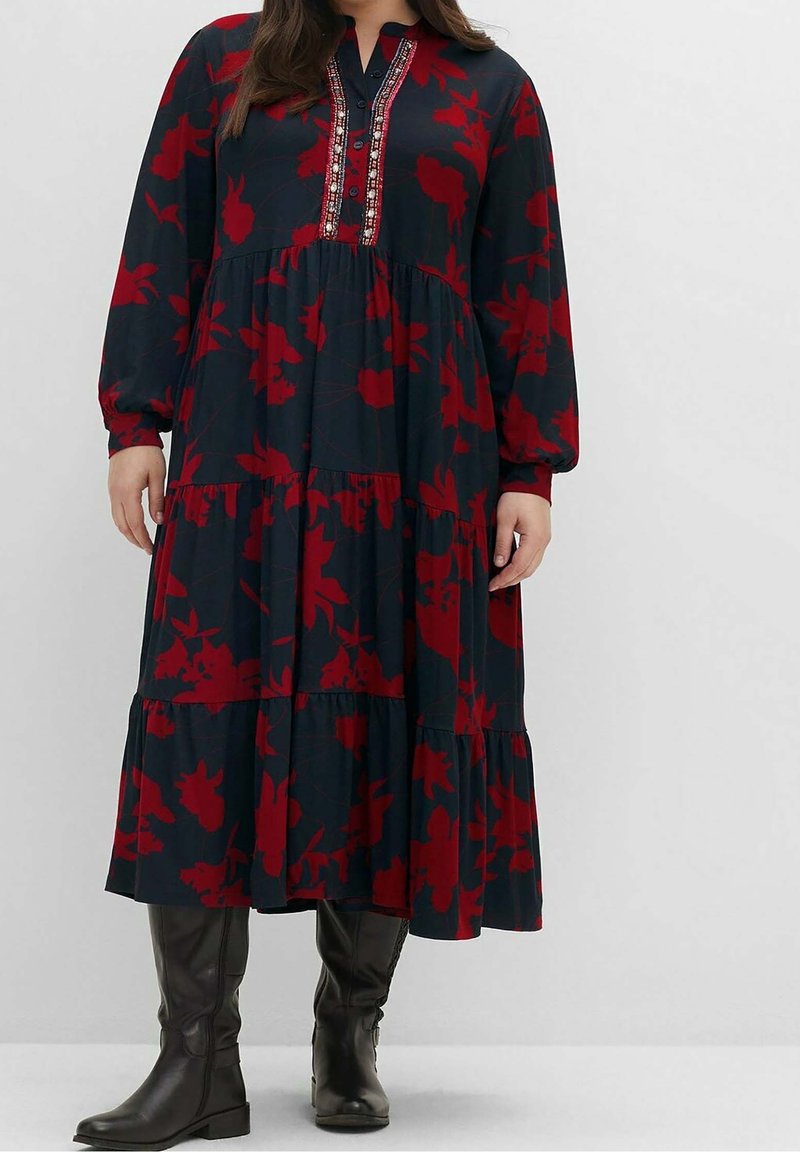Femme portant une robe midi bleu foncé avec imprimé floral rouge, manches longues, encolure boutonnée décorative, et bottes noires montantes jusqu'aux genoux.