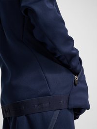 Person trägt eine marineblaue Sportjacke mit seitlicher Reißverschlusstasche und gebrandetem elastischem Taillenbund, seitlich vor neutralem Hintergrund gezeigt.
