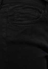 Κοντινή λήψη της τσέπης μαύρου τζιν με διακριτικό λογότυπο "A|X" κεντημένο στην μικρή μπροστινή τσέπη για κέρματα.