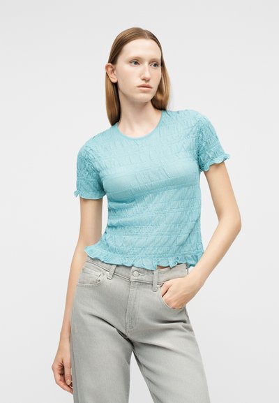 Γυναίκα με ίσια ανοιχτό καφέ μαλλιά, φοράει μπλε ανοιχτό κοντομάνικη μπλούζα με υφή και ανοιχτό γκρι τζιν, με το ένα χέρι στην τσέπη.
