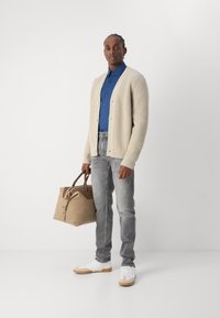 Cardigan beige acanalado, camisa azul de botones, jeans grises, zapatillas blancas con suelas tostadas, sosteniendo un bolso tostado con acentos de cuero marrón.