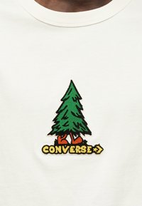 Άσπρο T-shirt με κεντητό πράσινο σχέδιο δέντρου με κόκκινα παπούτσια και κίτρινο κείμενο "CONVERSE" από κάτω. Απλό, καθημερινό στυλ.