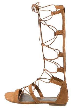 Sandalia gladiador marrón hasta la rodilla con tiras delgadas y cierre de cordones, suela plana y diseño con punta abierta.