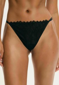 Kobieta w czarnych koronkowych majtkach w stylu bikini o niskim stanie, pokazująca środkową część ciała i górną część ud, z rękami luźno opadającymi wzdłuż ciała.