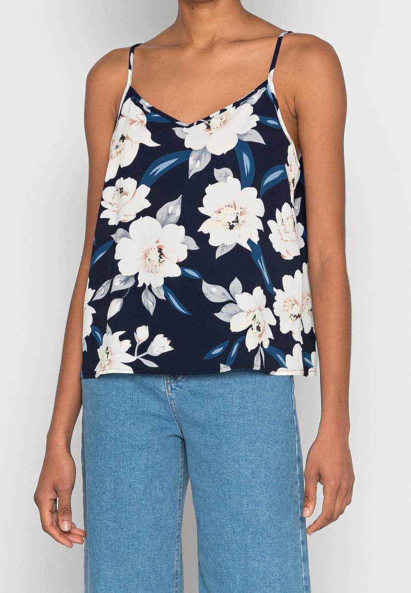 Femme portant un haut bleu marine à fines bretelles avec imprimé floral blanc et un jean taille haute bleu clair, debout, bras détendus.