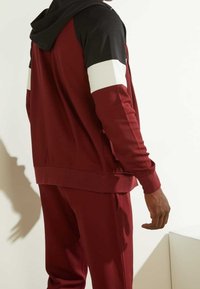 Chándal deportivo granate con chaqueta con capucha y cremallera, con mangas acentuadas en negro y blanco, tela suave y corte holgado.