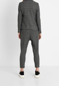 Szara kraciasta marynarka i spodnie zwężane, z kontrastującymi białymi paskami na mankietach, noszone z czarnymi tenisówkami na białym tle.