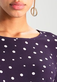 Top viola con pois bianchi, caratterizzato da un collo rotondo e una texture liscia. Un grande orecchino circolare d'oro mette in risalto il design.