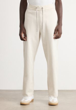 Person wearing beige drawstring pants and white sneakers, standing on a light floor against a plain white background.