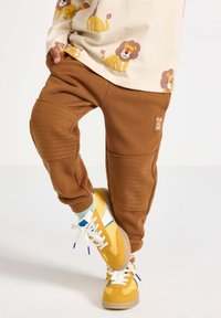 Braune Sweatpants mit gerippten Knie-Akzenten und einem Patch "PLAY ALL DAY", kombiniert mit gelben Sneakers mit weißen und blauen Details.