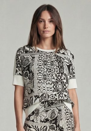 Woman wearing a black and white short-sleeve top and skirt with intricate floral and geometric patterns against a plain gray background.