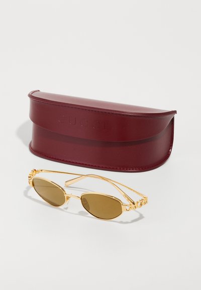 Kultaiset kehykset, ovaalimaiset aurinkolasit ruskeilla linsseillä ja Gucci-logoilla sangan päällä, asetettuna viininpunaisen kaarevan kovakotelon viereen, jossa on Gucci-logo.