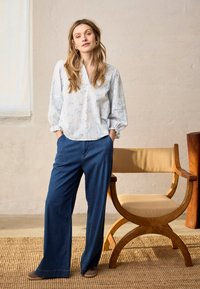 Jeune femme aux longs cheveux, les mains dans les poches, portant un chemisier léger à motifs, un large pantalon bleu et des chaussures marron, debout à côté d'une chaise en bois.