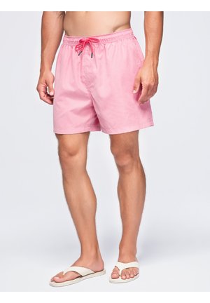 Homme portant un short de bain rose et des tongs blanches, debout devant un fond clair uni.