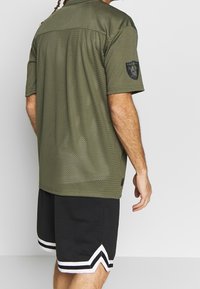 Man med olivgrön perforerad Raiders kortärmad tröja och svarta basketshorts med vit chevront trim, vänd bort.