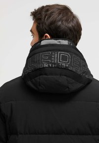 Schwarze Pufferjacke mit hohem Kragen, ausgestattet mit eingeprägtem Branding am Kragen. Der Stoff ist glatt und untexturiert, mit einem matten Finish.