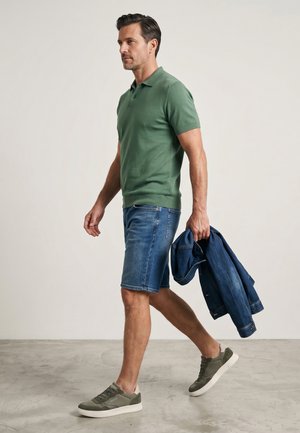 Muž chodí uvnitř, má na sobě zelené polo tričko, džínové kraťasy, šedé tenisky a v pravé ruce drží složenou modrou džínovou bundu.