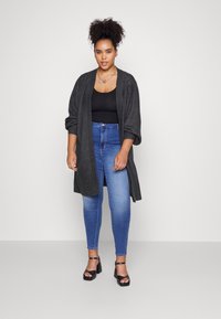 Gray oversized cardigan, black tank top, blue skinny jeans, and black heeled sandals; model stands against a neutral background.