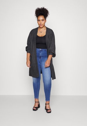 Grauer übergroßer Cardigan, schwarzes Tanktop, blaue Skinny-Jeans und schwarze High Heels; das Model steht vor einem neutralen Hintergrund.