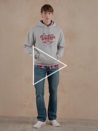 Sudadera con capucha gris con gráfico rojo, camisa de cuadros azul debajo, vaqueros azules y zapatillas blancas con detalles rojos. De pie sobre un fondo texturizado.
