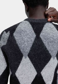 Persona che indossa un maglione a rombi nero e grigio, visibile di spalle, sta sistemando il colletto con la mano destra.