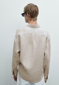 Beige Leinenhemd mit Kragen, Rückennaht und mit Knöpfen versehenen Manschetten. Modell trägt Sonnenbrille, von hinten betrachtet vor einem neutralen Hintergrund.