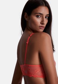 Soutien-gorge en dentelle corail avec bretelles réglables, détails de broderie florale et dos transparent. Texture lisse et accents de couture délicates.