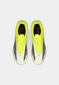 Ποδοσφαιρικά παπούτσια σε λαχανί πράσινο με υφή λευκού επάνω μέρους, μαύρες ρίγες και κίτρινα κορδόνια. Διαθέτουν κόκκινο λογότυπο και επένδυση στον γιακά για άνεση.