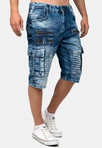 Shorts in denim con un lavaggio blu sbiadito, con diverse tasche con zip e pannelli testurizzati. Presentano una vestibilità rilassata e un design fino al ginocchio.