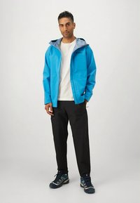 Kék vízálló dzseki szürke kapucnival, fekete nadrággal és kék tornacipővel párosítva. A dzsekin cipzár az elején és logó részlet található.