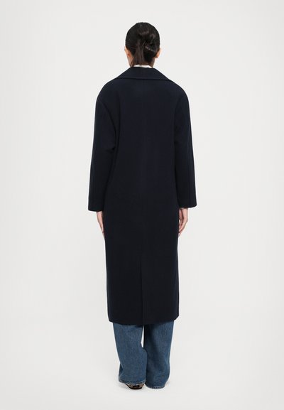 Donna dai capelli scuri raccolti in un bun, che indossa un lungo cappotto scuro e jeans blu, in piedi di spalle contro uno sfondo bianco semplice.