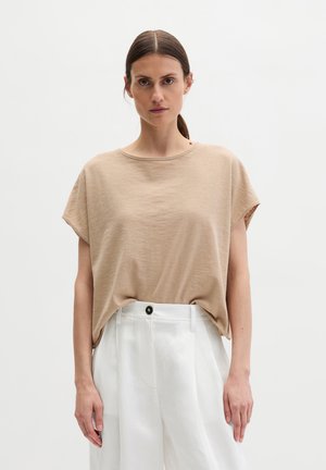 Femme aux cheveux bruns attachés, portant un haut beige ample à manches courtes et un pantalon blanc taille haute, sur un fond blanc uni.