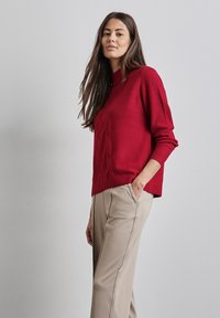 Maglione rosso lavorato a maglia con texture a costine e collo rotondo, abbinato a pantaloni beige con tasche laterali e cuciture in evidenza.